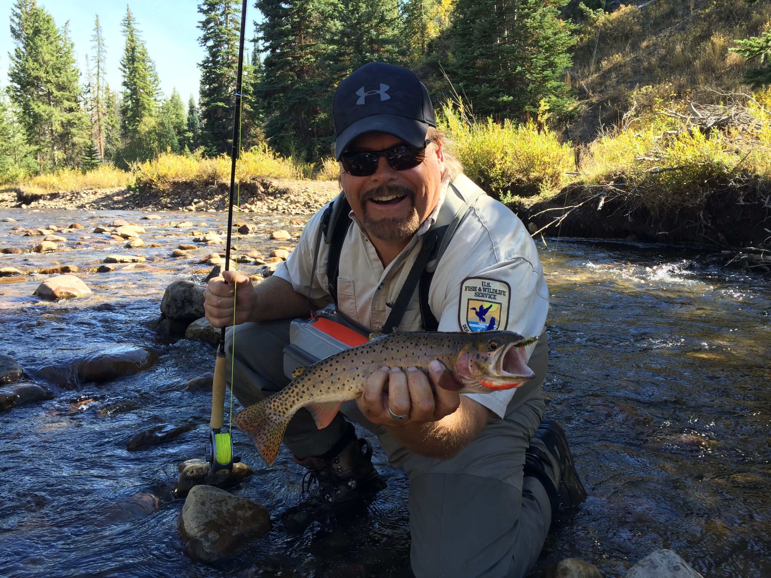 A U.S. Fish and Wildlife Service employee holds a fish and a fly rod while crouching in a river.