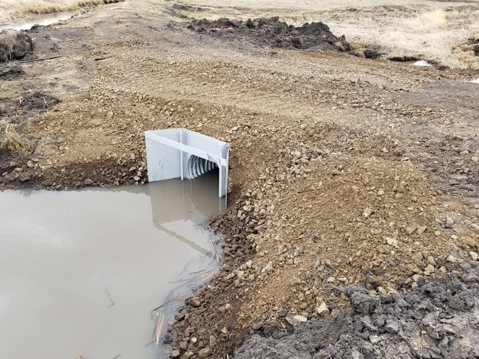 A new culvert with flashboard risers runs under a dirt road.