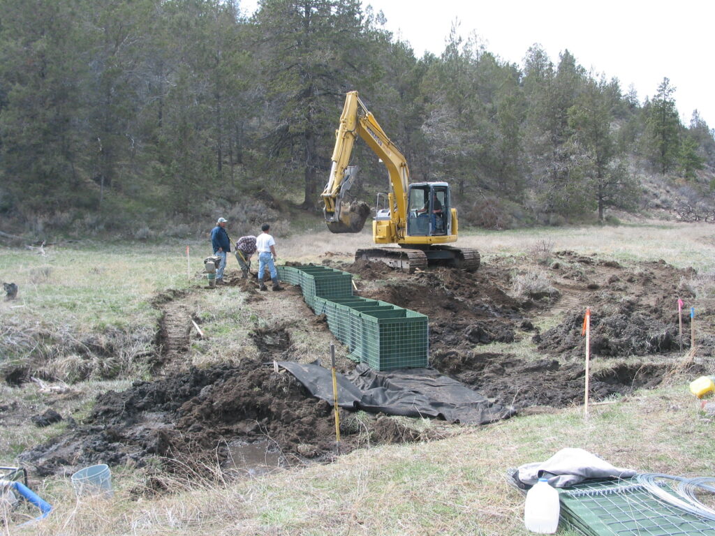 A backhoe puts fill into green boxes as people watch.