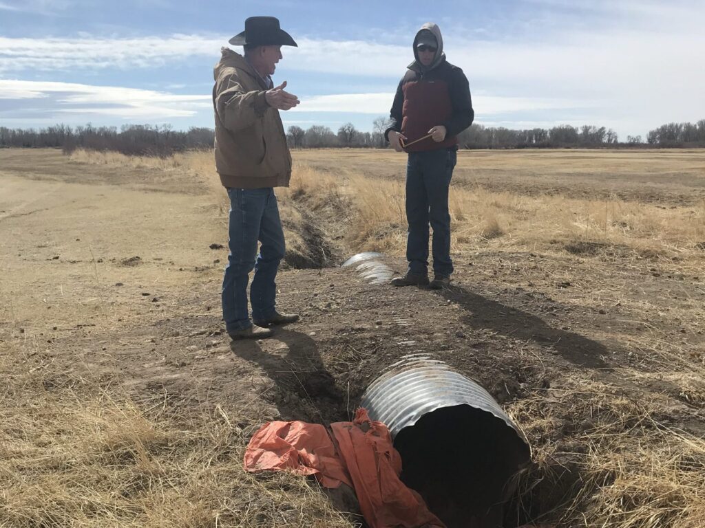 Two people talk over an old culvert in a dry hay field.
