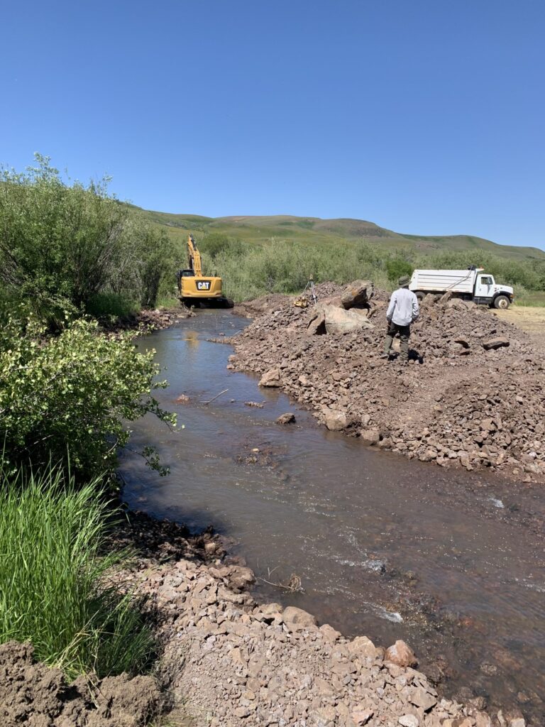 Machinery in a creek and a man on a pile of dirt on its banks.
