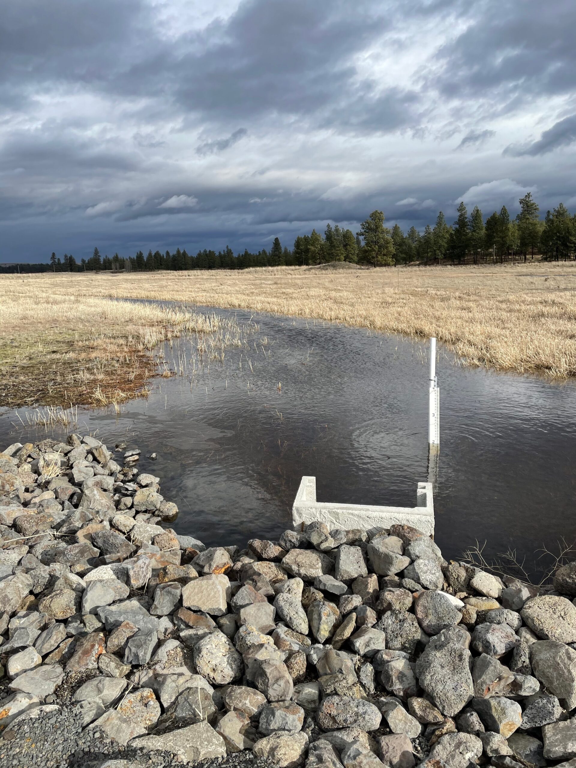 Rip-rap, a measuring device, and a new concrete culvert in a flooded field.